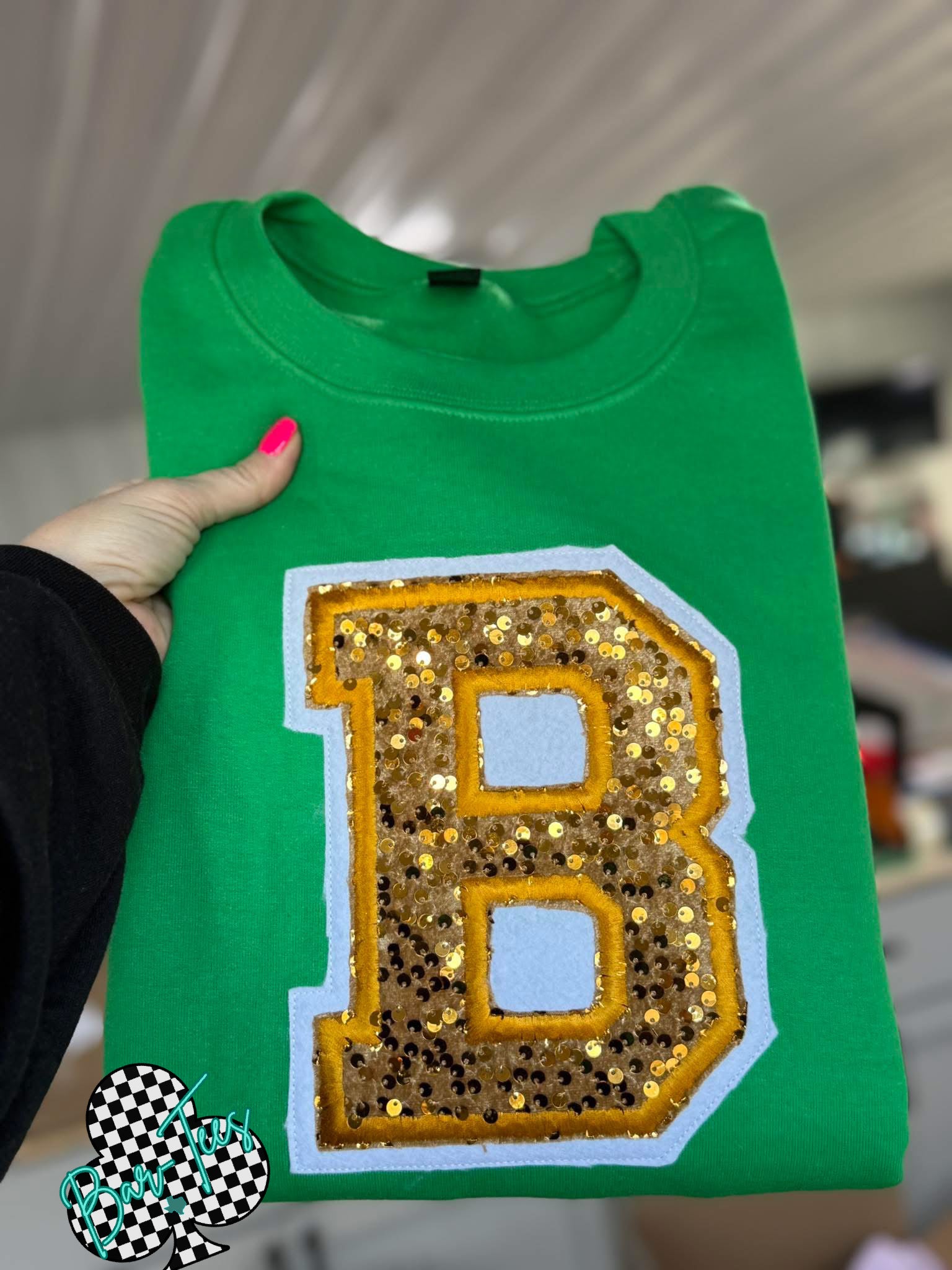 Green sweatshirt with a large sequined letter 'B' held by a person.