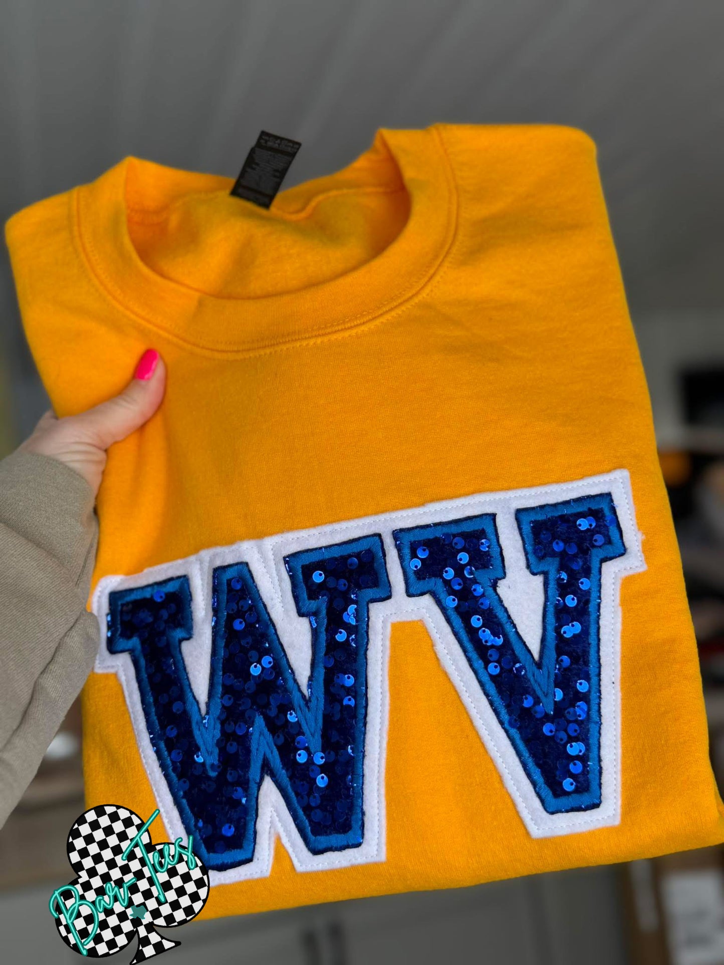 Yellow sweatshirt with blue 'WV' letters held by a person against a blurred indoor background.